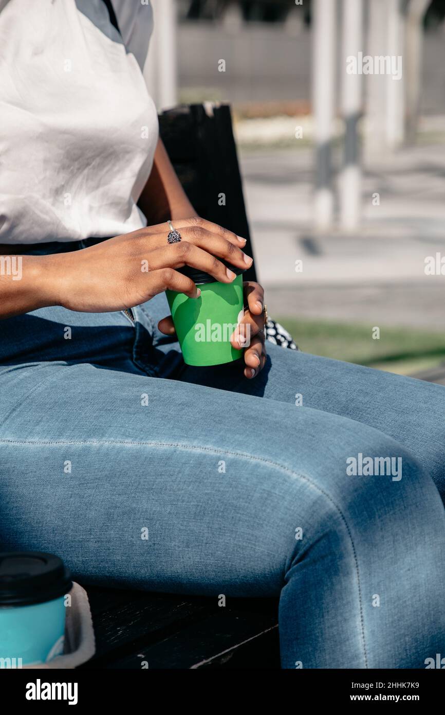 Crop African American weiblich in legerer Kleidung sitzt auf der Bank mit einer Tasse Kaffee zum Mitnehmen an sonnigen Tag in der Stadt Stockfoto