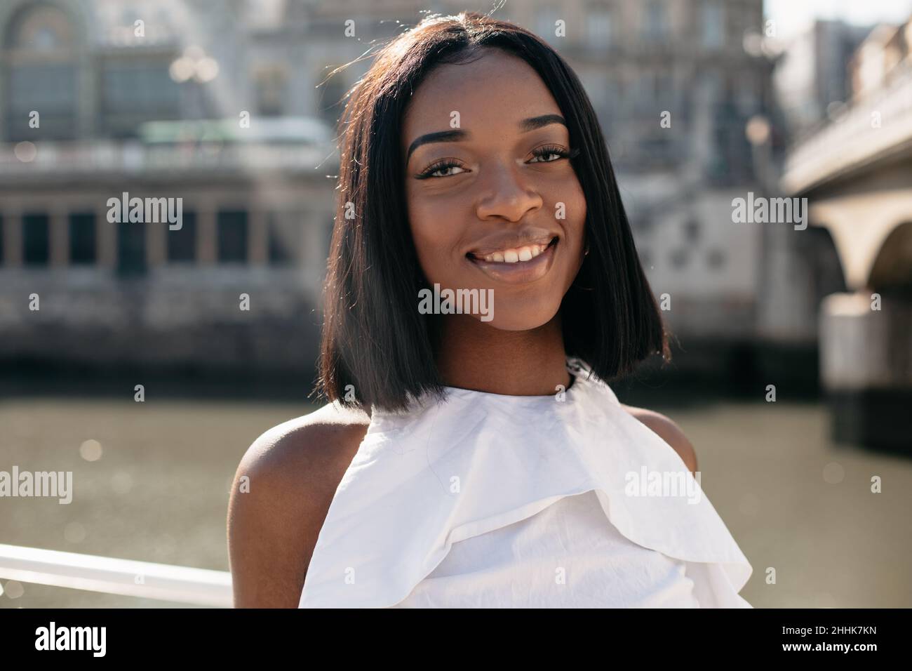 Glückliche afroamerikanische Frau mit kurzen, geraden Haaren, die lächelte und die Kamera betrachte, während sie tagsüber auf der sonnenbeschienenen Stadtstraße stand Stockfoto