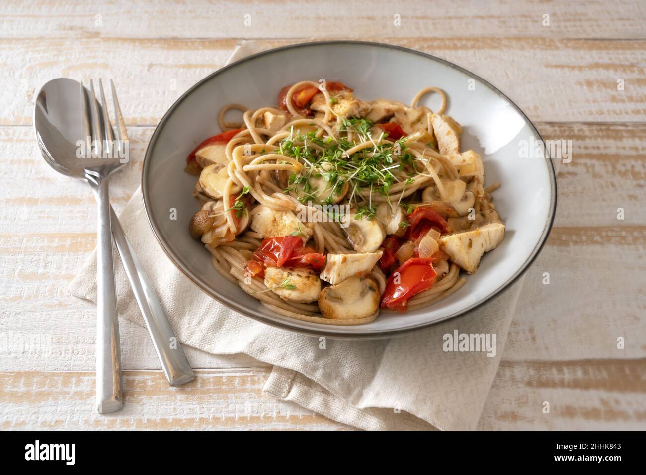 Vollkorn-Spaghetti mit Tomaten, Pilzen und Hähnchenstreifen, garniert mit Kresse, grauem Teller und Besteck auf einem hellen rustikalen Holztisch, ausgewählt Stockfoto