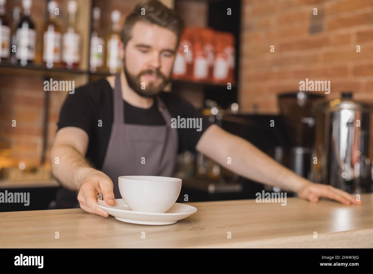 Bärtiger Barista auf der Schürze, der eine Tasse Kaffee serviert, während er am Bartisch im Café in der Nähe der Kaffeemaschine arbeitet Stockfoto
