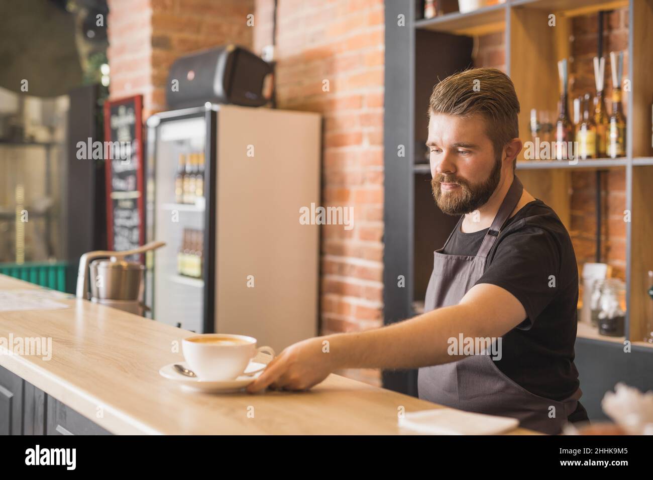 Bärtiger Barista auf der Schürze, der eine Tasse Kaffee serviert, während er am Bartisch im Café arbeitet Stockfoto