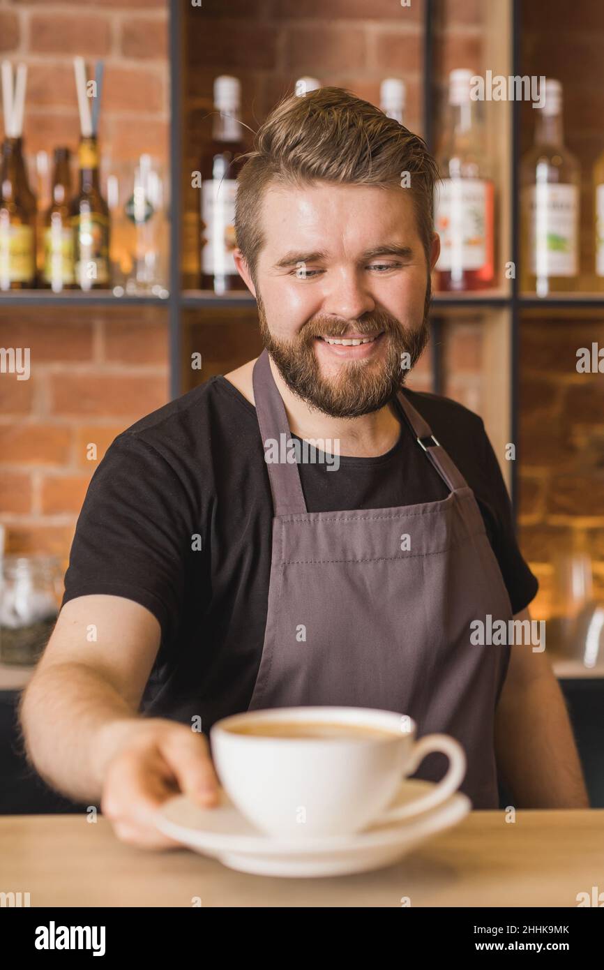 Bärtiger Barista auf der Schürze, der eine Tasse Kaffee serviert, während er am Bartisch im Café arbeitet Stockfoto