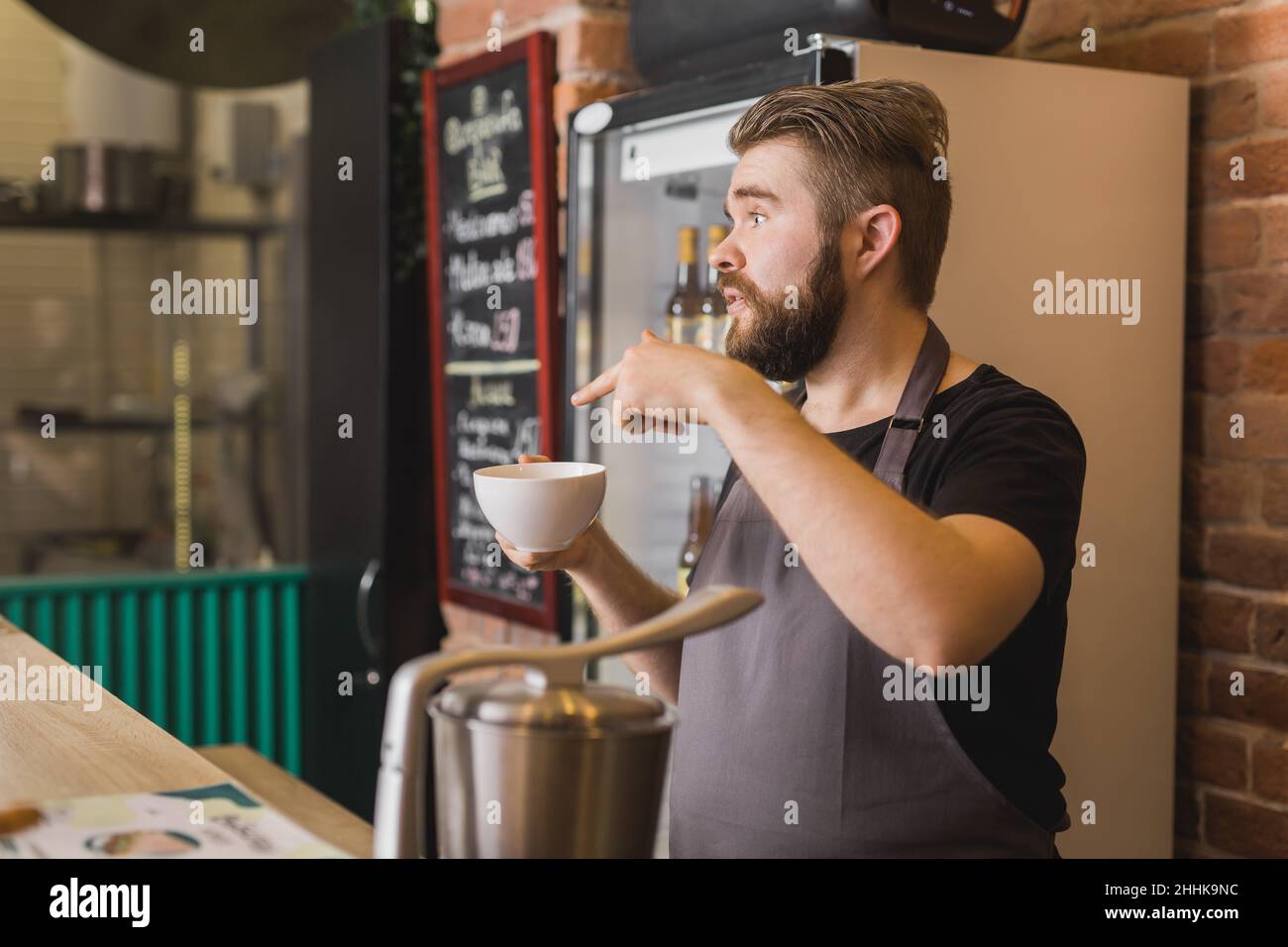 Seitenansicht des bärtigen männlichen Barista im Vorfeld, der mit einer Tasse heißem Getränk am Bartisch stand und dem Gast im Café die Bestellung gab Stockfoto