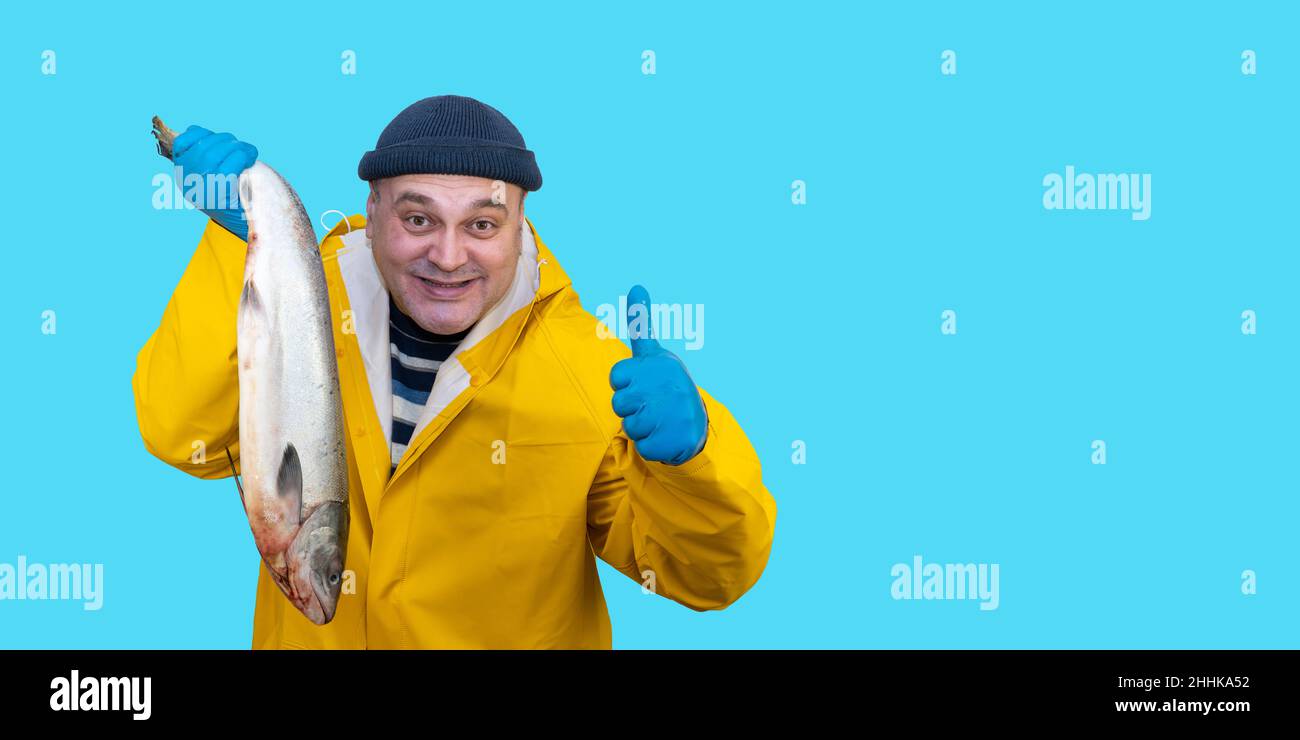 Fischer im gelben Anzug hält einen frisch gefangenen Fisch in den Händen. Fänger im Regenmantel. fischer auf blauem Hintergrund Stockfoto