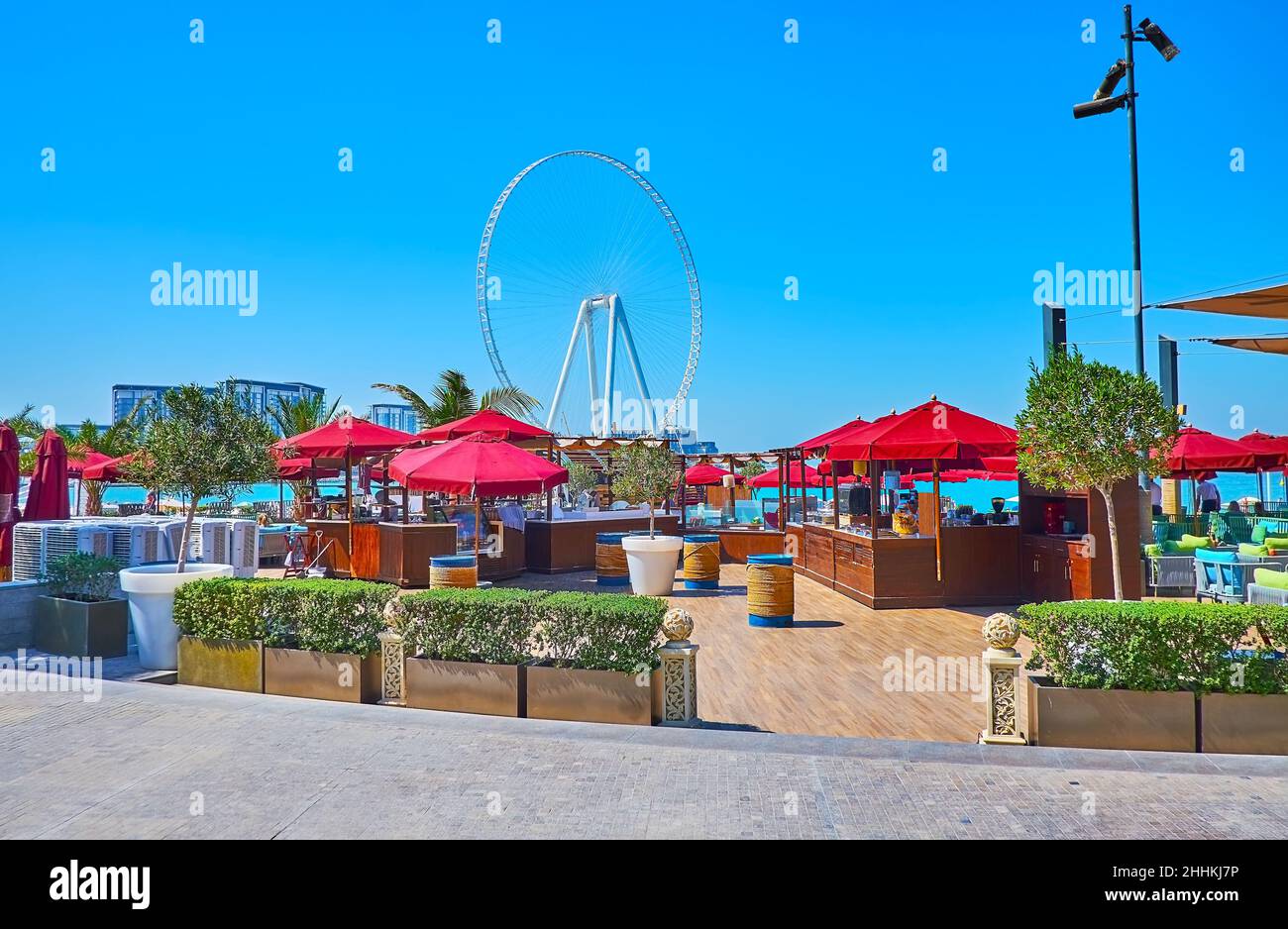 Die gemütliche Terrasse des Strandrestaurants mit roten Sonnenschirmen, Pflanzen in Töpfen und Blick auf das Meer und Ain Dubai Riesenrad, JBR Marina Beach, Dubai, Stockfoto