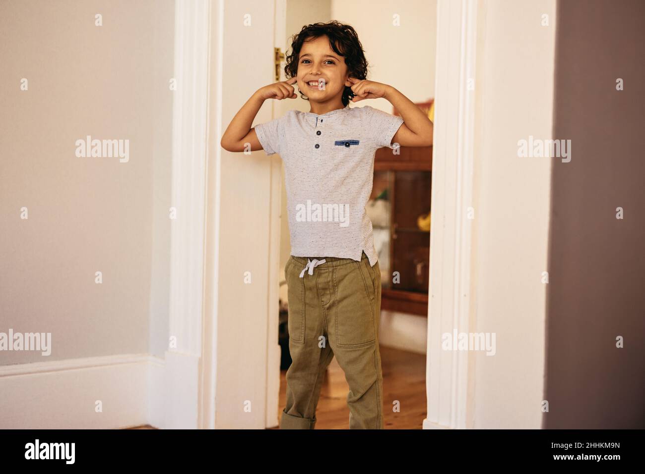 Glücklicher Junge, der zu Hause spielerisch seine Ohren bedeckt. Unbeschwerter ethnischer Junge, der fröhlich lächelt, während er im Haus an einer Tür steht. Stockfoto