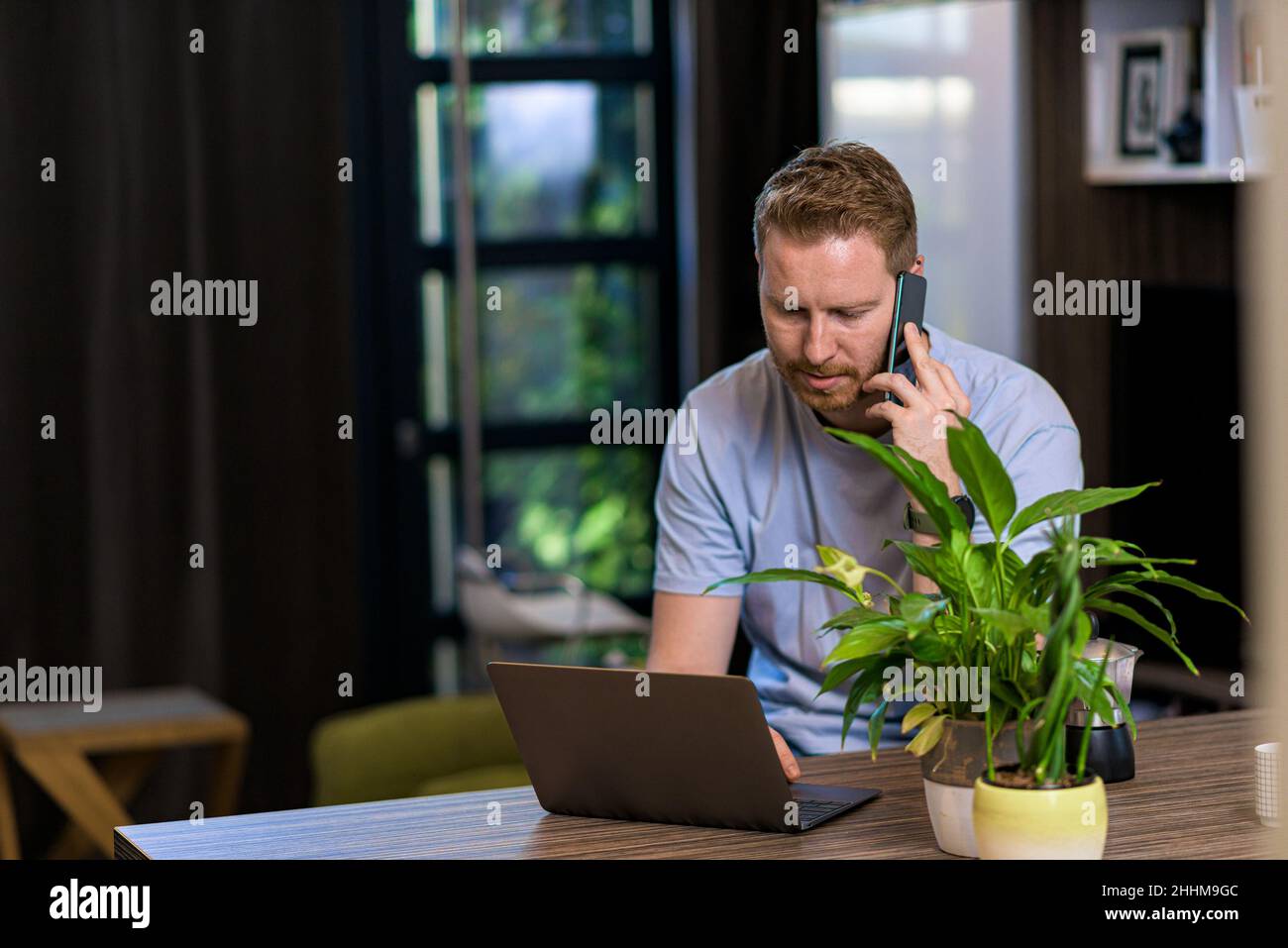 Porträt eines zwanglosen, nachdenklichen, rätselhaften Mannes, der ein Smartphone verwendet und einen Laptop im Heimbüro ansieht, der Probleme beim Anruf beim Kundendienst löst Stockfoto