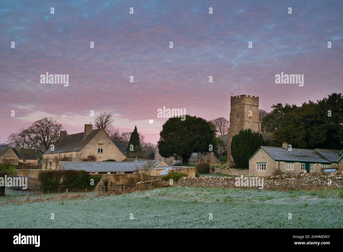 Blick über Hampnet Dorf im Winter Frost in der Morgendämmerung mit dem Vollmond und rosa Wolken am Himmel. Hampnet, Cotswolds, Gloucestershire, Großbritannien Stockfoto