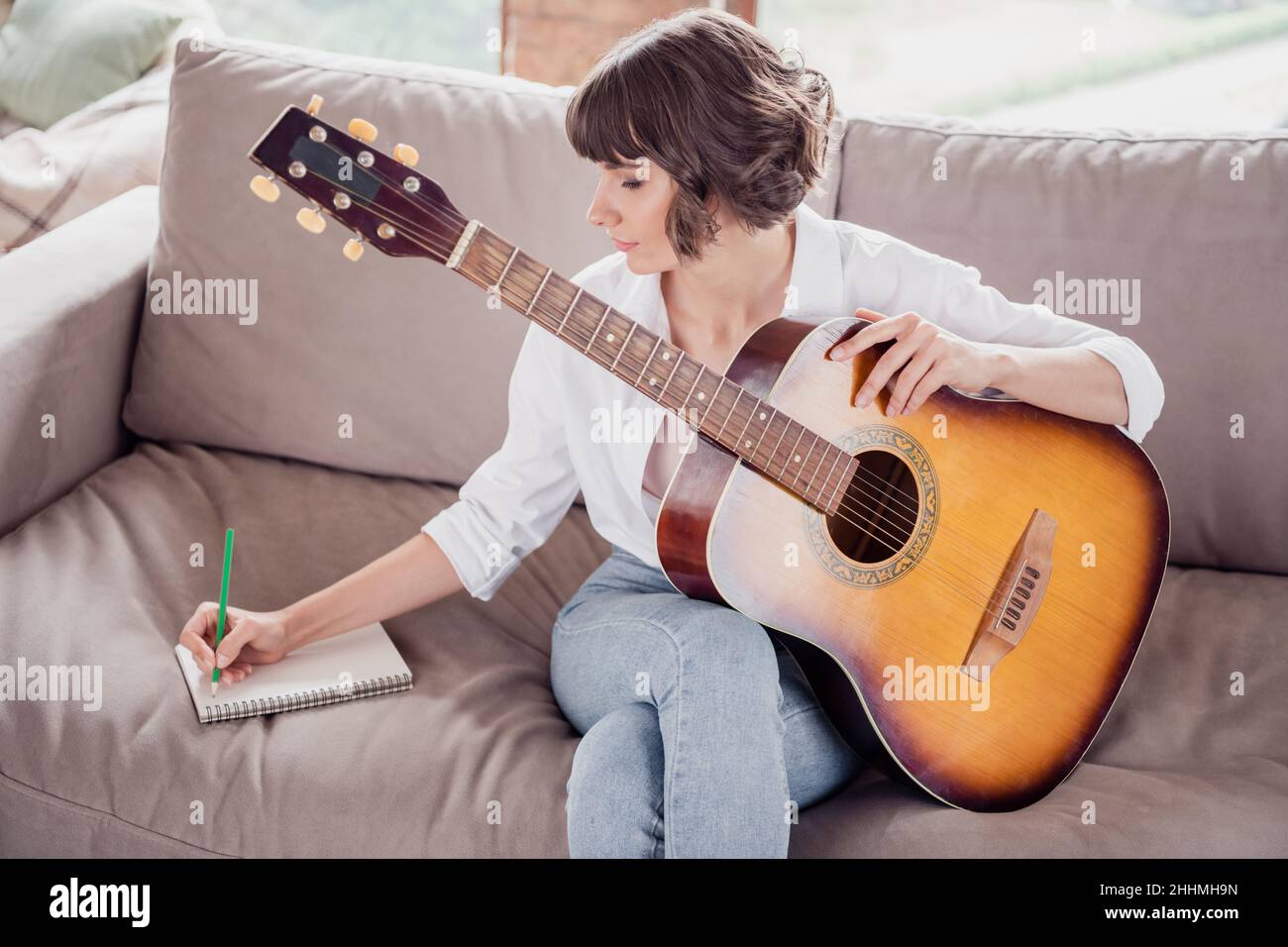 Portrait Foto junge Frau, die lernt, akustische Gitarre zu spielen, sitzt auf dem Sofa und schreibt Notizen mit Bleistift Stockfoto