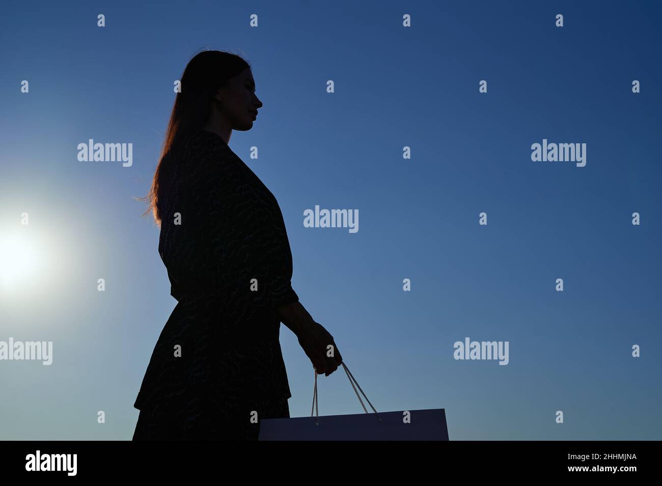 Silhouette der kaukasischen modischen Frau mit langen Haaren Blick auf Einkaufstasche einen Sonnenuntergang, Bild gegen Sonne und blauen Himmel. Konsumdenken, Shopping, Verkauf oder Black friday Konzept. Hochwertige Fotos Stockfoto