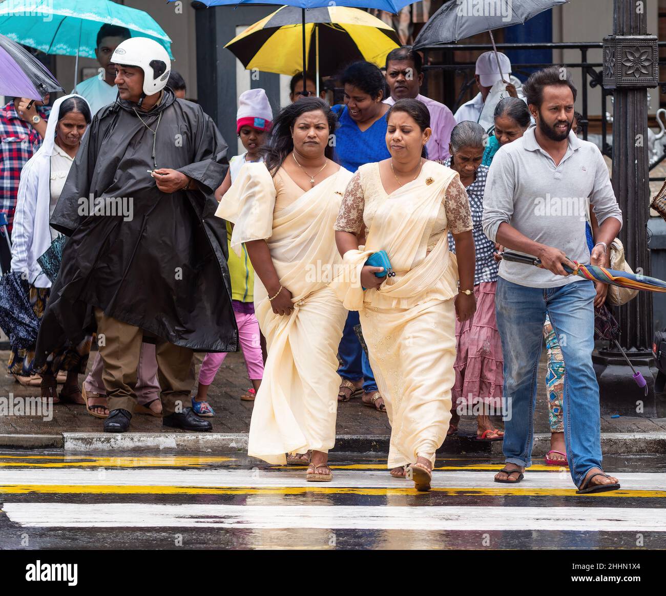 Menschen, die an einem Zebrastreifen an einem regnerischen Tag in Kandy, Sri Lanka, die Straße überqueren. Zwei Frauen im traditionellen Kandyan Saree aufgrund eines religiösen Festivals Stockfoto