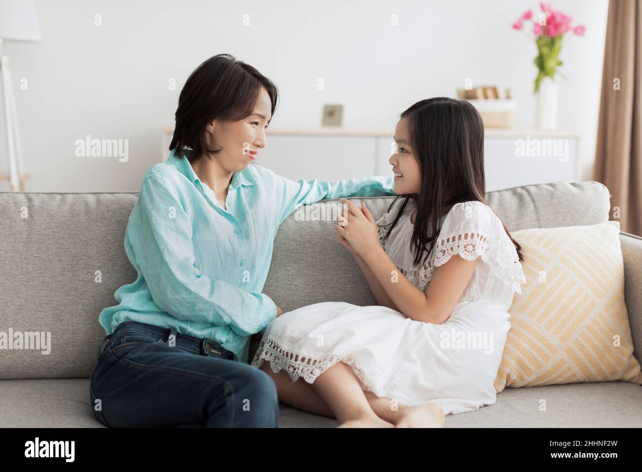 Glückliches kleines asiatisches Mädchen, das mit ihrer lächelnden Großmutter spricht und zu Hause auf der Couch sitzt Stockfoto