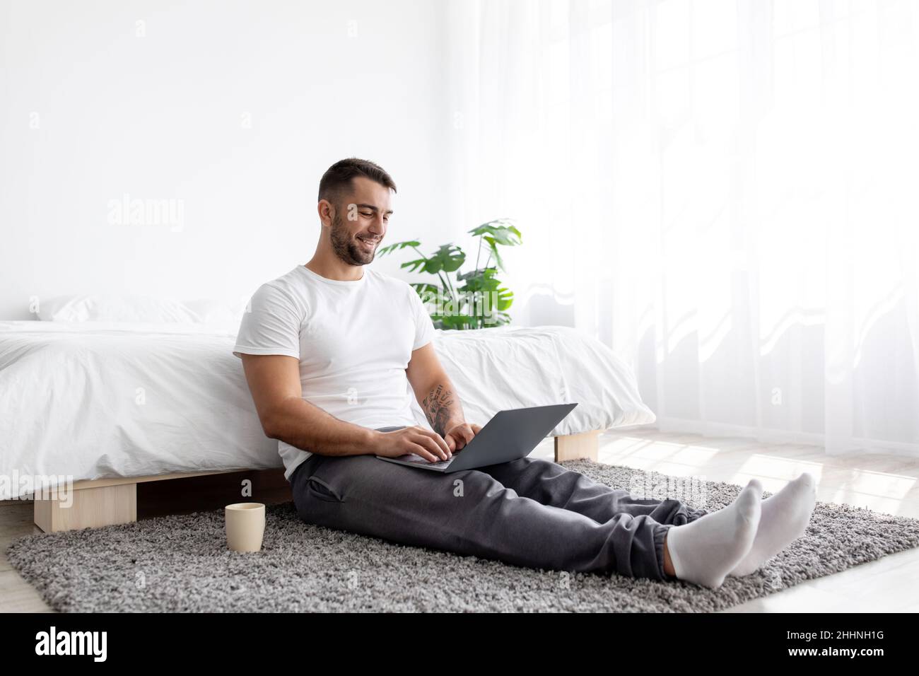 Lächelnder junger kaukasischer, hübscher, muskulöser Mann, der auf dem Boden sitzt und im Schlafzimmer auf dem Laptop tippt Stockfoto