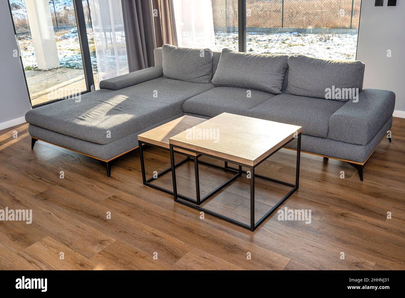 Ein modernes und einfaches Wohnzimmer im Haus mit einem grauen Sofa, sichtbaren großen Terrassenfenstern und zwei Tischen, die sich untereinander befinden. Stockfoto