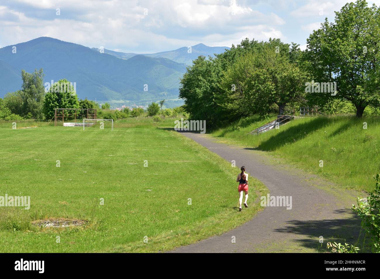 Junge Langstreckenläuferin in roten Shorts und schwarzem Sweatshirt läuft in einem Sportstadion mit grünem Gras. Stockfoto