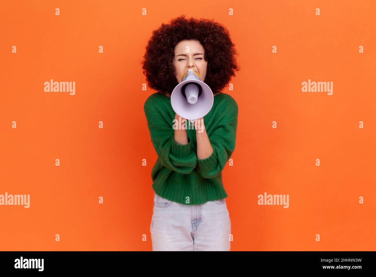 Attraktive Frau mit Afro-Frisur trägt grünen lässigen Stil Pullover schreiend laut mit Megaphon, die Ankündigung. Präsentation. Innenaufnahme des Studios isoliert auf orangefarbenem Hintergrund. Stockfoto