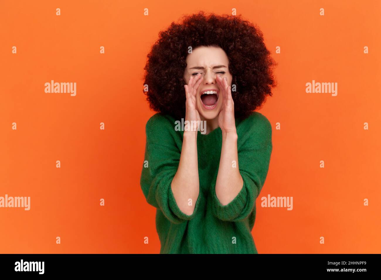 Porträt einer wütenden schreienden Frau mit Afro-Frisur, die einen grünen Pullover im lässigen Stil trägt, die Hände in der Nähe des Mundes hält und laut schreit. Innenaufnahme des Studios isoliert auf orangefarbenem Hintergrund. Stockfoto