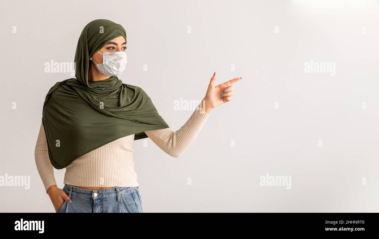 Coronavirus-Schutz. Junge arabische Frau in Hijab und antiviraler Gesichtsmaske, die auf leeren Raum auf hellem Hintergrund zeigt Stockfoto