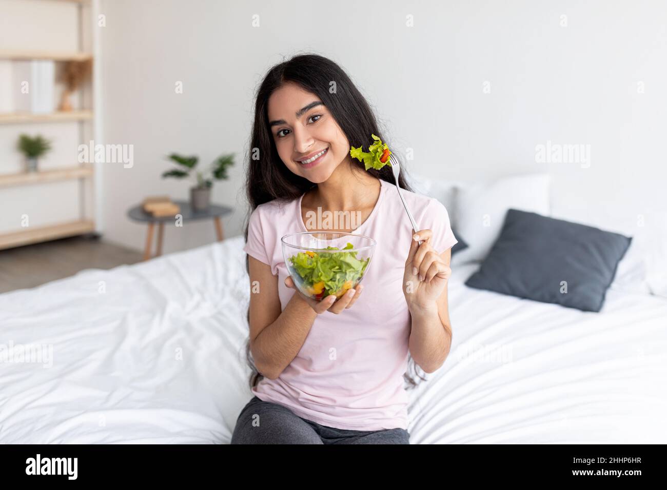 Positive indische Frau, die zu Hause auf dem Bett sitzt und leckeren Gemüsesalat isst. Gesunde Ernährung zur Gewichtsabnahme Konzept Stockfoto