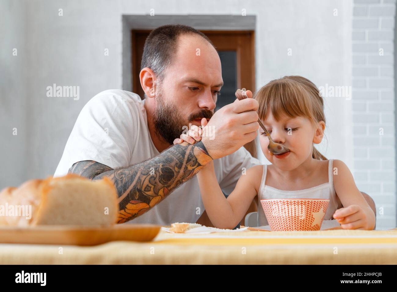 Vater füttert seine Tochter mit Löffel. Kahlbärtig tätowiert kaukasischen Mann Vater gibt Nahrung füttert sein Kind Kind Tochter sanfte blonde Mädchen mit Stockfoto