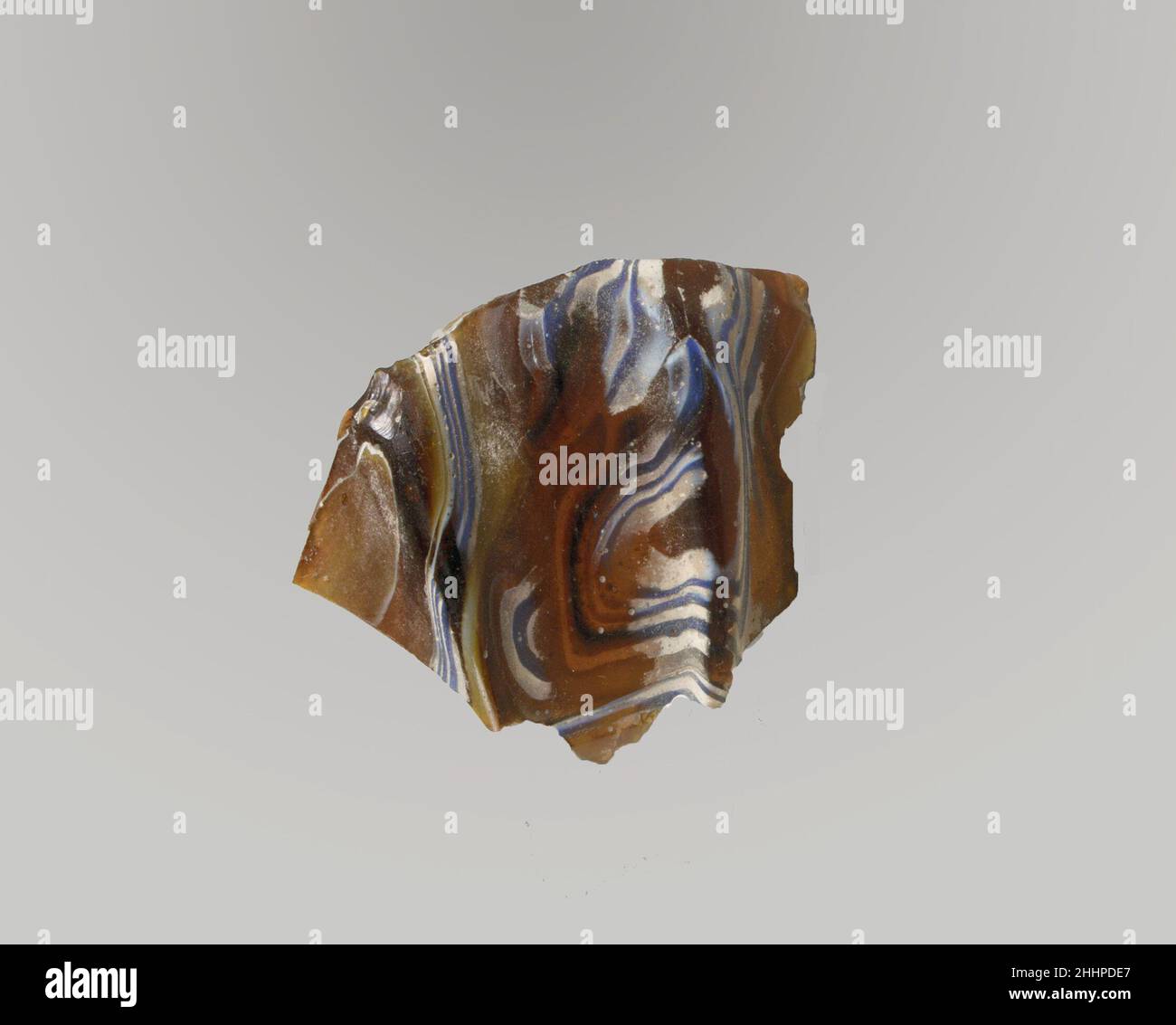 Glasmosaik geripptes Schalenfragment Ende des 1st. Jahrhunderts v. Chr.–Anfang des 1st. Jahrhunderts n. Chr. römisches Körperfragment. Lichtdurchlässiges honigbraun, kobaltblau und opakes weiß. Konvex geschwungene Seite, stark zugespitzt. Spiralmosaik-Muster aus Abschnitten eines einzelnen Rohres in braunem Boden mit unregelmäßigen weißen und blauen Fäden in parallelen Linien gebildet; Außen zwei senkrechte Rippen, mit abgeflachten Oberteilen und abgerundeten Außenkanten, nach unten verjüngend. Außen poliert; außen mit Lochfraß von Oberflächenblasen; außen stumpfend und cremig schillernd, außen mit gezackten Kanten. Glasmosaik gerippte Schale Fragment. Roman. Spät Stockfoto