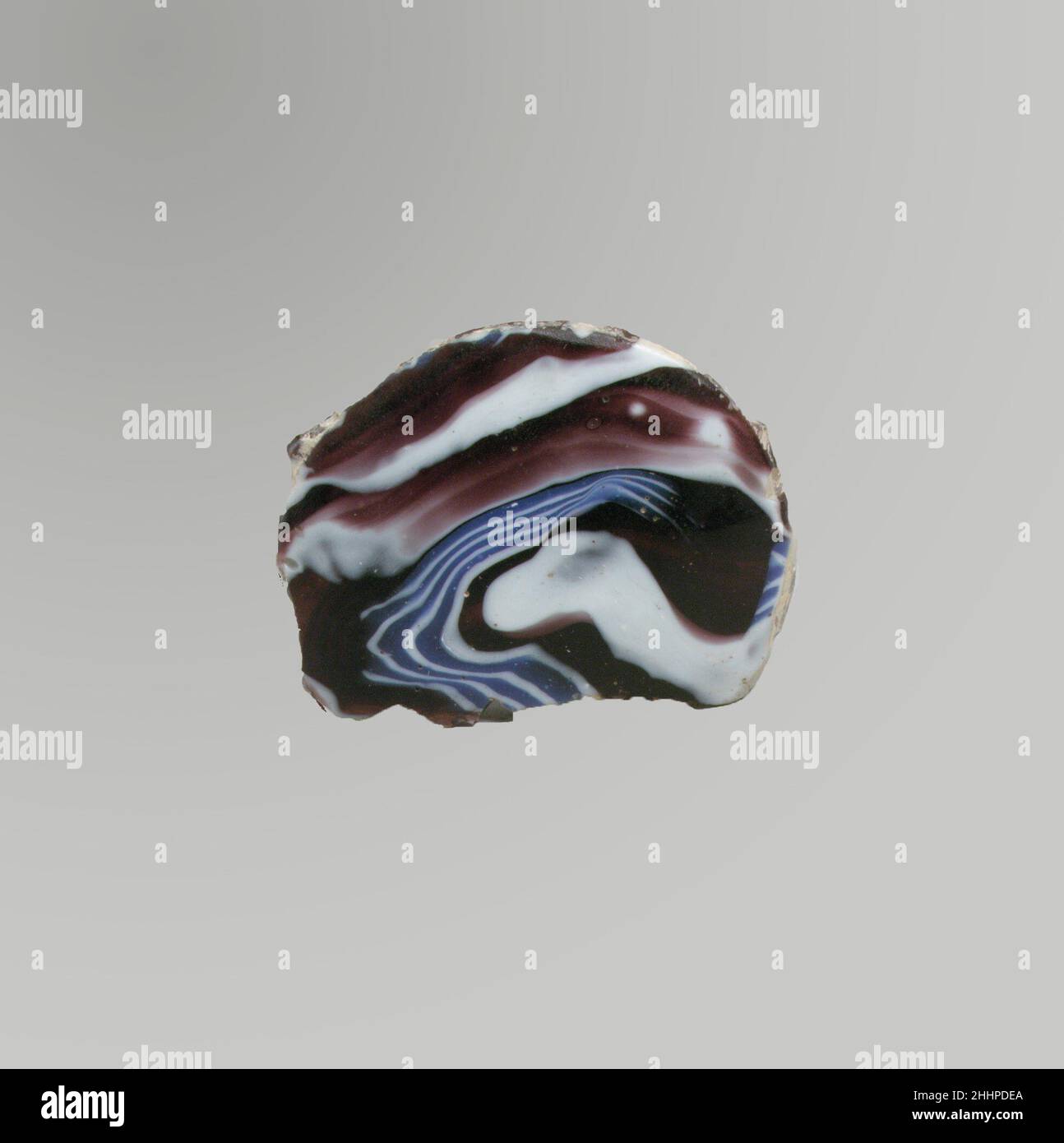 Glasmosaik-Schalenfragment Ende des 1st. Jahrhunderts v. Chr.–Anfang des 1st. Jahrhunderts n. Chr. römisches Bodenfragment.Durchscheinendes Lila, Kobaltblau und opakes Weiß.dicker gewölbter gewölbter Boden.Mosaikmuster aus Abschnitten eines einzelnen Rohres in einem violetten Boden mit unregelmäßigen weißen Spritzern und welligen weißen und blauen Fäden in parallelen Linien. Lochfraß von Oberflächenblasen auf der Außenseite; cremig schillernde Verwitterung bedeckt den Großteil des Innenraums; Schillern an den Kanten. Möglicherweise in der Antike in eine Scheibe umgearbeitet.wahrscheinlich Teil der konkaven Basis einer gerippten Schale.. Fragment der Glasmosaik-Schale. Roman, l Stockfoto