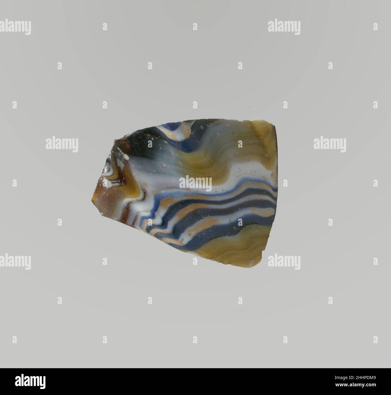 Glasmosaik gerippte Schale Fragment Ende 1st Jahrhundert v. Chr.–Anfang 1st Jahrhundert n. Chr. Römisches Körperfragment. Lichtdurchlässig honigbraun, teilweise mit Weiß gemischt und undurchsichtig gelb, blau, Und opakes Weiß.konvex geschwungene Seite, nach unten zulaufend.Mosaikmuster mit Band, das aus Längen eines einzelnen Rohres in einem braunen Boden mit Zickzack-Bändern aus weißen und blauen Fäden in parallelen Linien und einfachen Bändern in Gelb gebildet wird; an der Außenseite untere Abschnitte von drei vertikalen Rippen, flach zulaufend.Polierte Außenseite; Lochfraß von Oberflächenblasen auf der Außenseite; stumpfende und cremig schillernde Verwitterung auf der Innenseite und Stockfoto