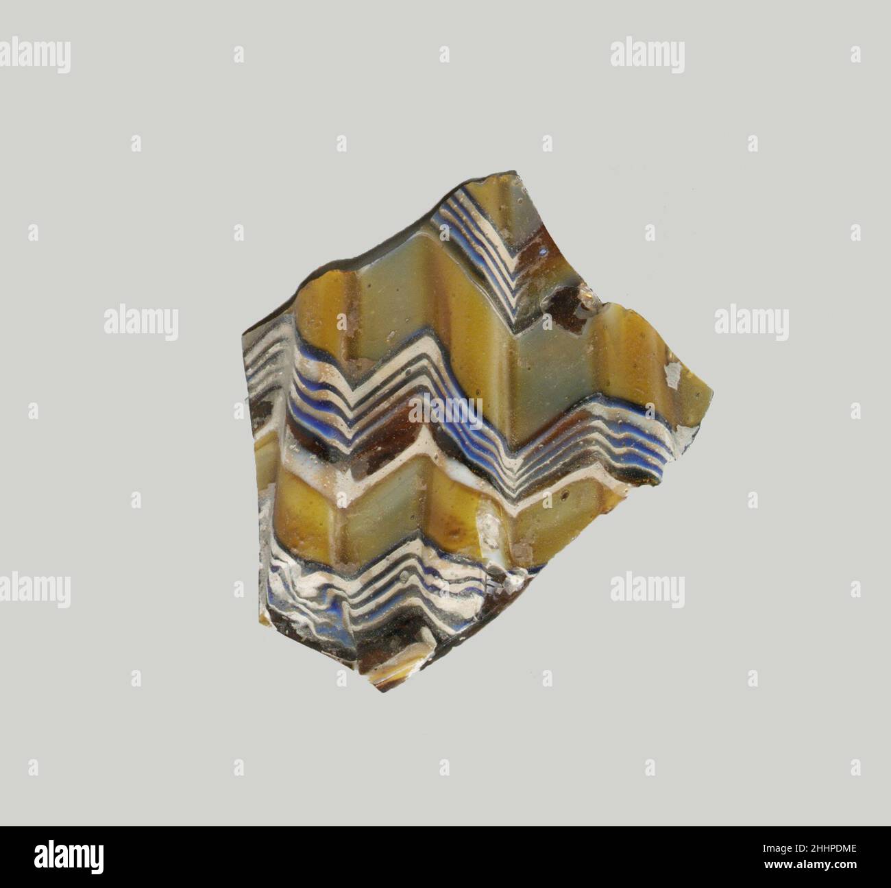 Glasmosaik gerippte Schale Fragment Ende 1st Jahrhundert v. Chr.–Anfang 1st Jahrhundert n. Chr. Römisches Körperfragment. Lichtdurchlässig honigbraun, teilweise mit Weiß gemischt und undurchsichtig gelb, blau, Und opakes Weiß.Fast gerade Seite, nach unten zulaufend.Mosaikband aus Längen eines einzelnen Rohres in einem braunen Boden mit Zickzack-Bändern aus weißen und blauen Fäden in parallelen Linien und einfachen Bändern in Gelb gebildet; Außen Teile von drei vertikalen Rippen, mit abgeflachten Oberteilen und abgerundeten Außenkanten. Außen poliert; außen mit Lochfraß von Oberflächenblasen und einigen verwitterten Spänen; außen stumpfend und cremig i Stockfoto