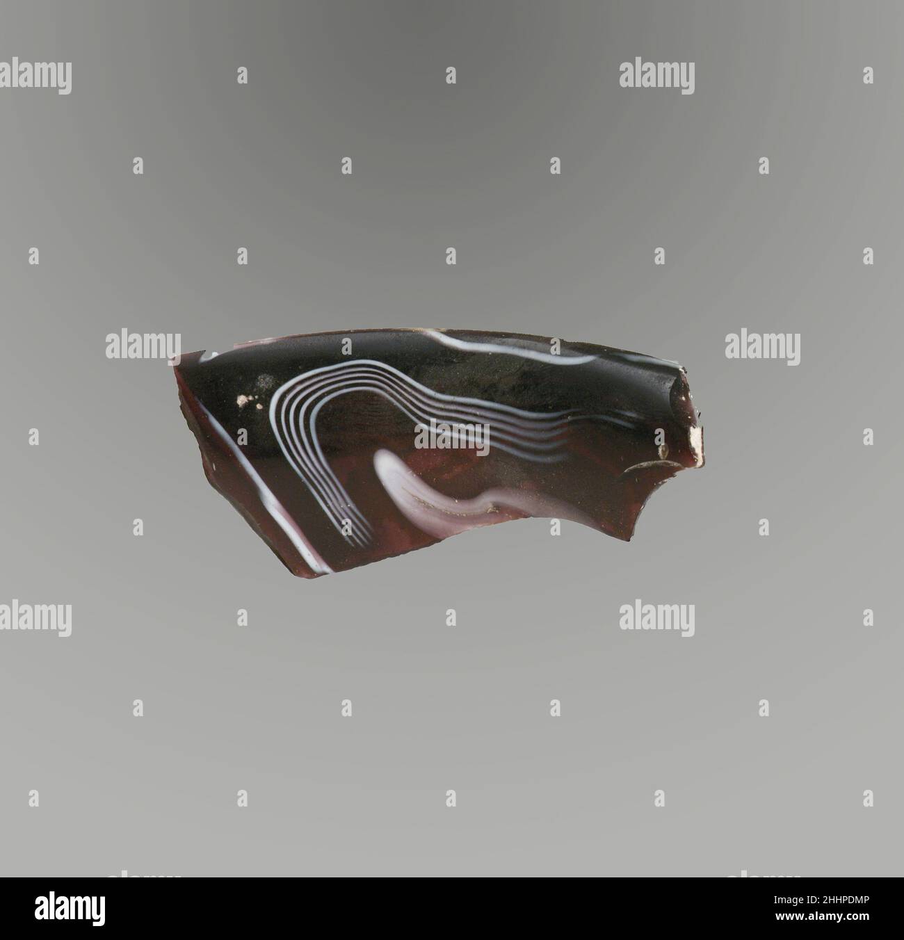 Glasmosaik gerippte Schale Fragment Ende 1st Jahrhundert v. Chr.–Anfang 1st Jahrhundert n. Chr. Roman Durchscheinend lila und opak weiß.dicker, fast horizontaler Rand mit abgerundeter Kante; Gerade Seite, leicht nach unten zulaufend.spiralförmiges Mosaikmuster, das aus einer oder mehreren Rohrstöcken in violettem Boden mit weißen Spiralen und feinen Fäden in parallelen Linien gebildet wird; außen Spuren einer vertikalen flachen Rippe.Poliertes Innere; Lochfraß von Oberflächenblasen; Stumpfend, cremig verwittert und etwas schillernd auf der Außenseite und gezackten Kanten. Glasmosaik gerippte Schale Fragment. Römisch. Ende des 1st. Jahrhunderts v. Chr.–Anfang des 1st. Jahrhunderts Stockfoto