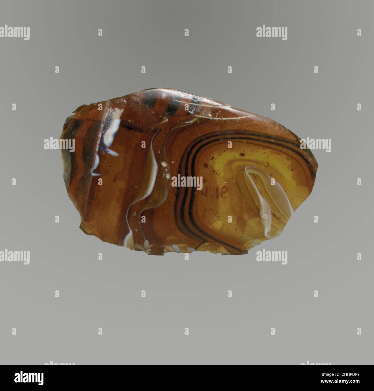 Glasmosaik-Schalenfragment Ende des 1st. Jahrhunderts v. Chr.–Anfang des 1st. Jahrhunderts n. Chr. Römisches dünnwandiges Körperfragment. Lichtdurchlässiges honigbraunes, blaues und opakes Weiß. Geschwungene Seite. Spiralförmiges Mosaikmuster, das aus Längen eines oder mehrerer Stöcke in einem braunen Boden mit welligen weißen und blauen Fäden in parallelen Linien gebildet wird; Außen eingelassenes horizontales Band.Pinprick-Blasen; polierte Außenseite; Lochfraß von Oberflächenblasen auf der Außenseite; stumpfende und cremig schillernde Verwitterung auf der Innenseite und gezackte Kanten. Fragment der Glasmosaik-Schale. Römisch. Spätes 1st Jahrhundert B.C.–frühes 1st Jahrhundert A.D.. Glas; gegossen und geschnitten. Früh Stockfoto