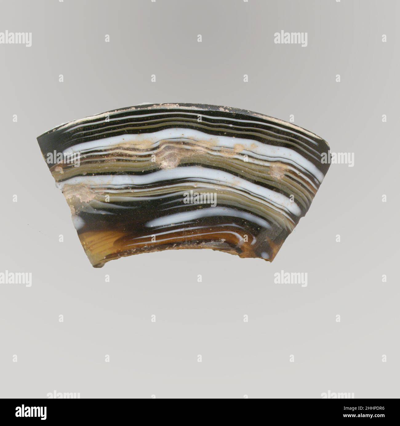 Glasmosaik-Schalenfragment Ende des 1st. Jahrhunderts v. Chr.–Anfang des 1st. Jahrhunderts n. Chr. römisch Transluzent honigbraun, tiefviolett schwarz und opak weiß.hoher, ausgespieleter Basisring, mit gewölbtem gewölbtem Rand im Inneren und abgerundetem unteren Rand; Unregelmäßige kleine kreisförmige Einbuchtungen an der Außenseite.Mosaikmuster aus einer oder mehreren Stöcken in violettem und braunem Grund mit feinen weißen Fäden, teilweise gelb, in parallelen Linien, zwischen breiten weißen Bändern angeordnet.Außen poliert; Lochfraß von Oberflächenblasen und Verwitterung in Einbuchtungen an der Außenseite; Cremige Verwitterung und etwas schillernd Stockfoto