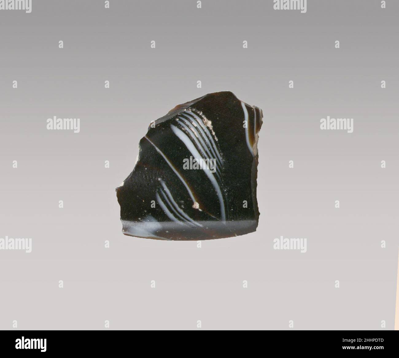 Glasmosaikfragment Ende des 1st. Jahrhunderts v. Chr.–Anfang des 1st. Jahrhunderts n. Chr. römisches Basisringfragment, wahrscheinlich aus einer tiefen, mit Füßen gerippten Schale.Durchscheinend tiefes Honigbraun und opakes Weiß.hoher, ausgespielter Basisring mit dicker, abgerundeter Kante; Konkave, kurvige Seite, die stark nach oben steigt.Mosaikmuster aus Bändern, die aus Abschnitten eines einzelnen Rohres in braunem Boden mit unregelmäßig welligen weißen Fäden in parallelen Linien gebildet werden.Poliertes Innere; Lochfraß von Oberflächenblasen im Inneren; stumpfende und cremig schillernde Verwitterung an der Außenseite, Rand der Basis und gezackte Kanten. Glasmosaikfragment. Römisch. Ende des 1st. Jahrhunderts v. Chr. Stockfoto