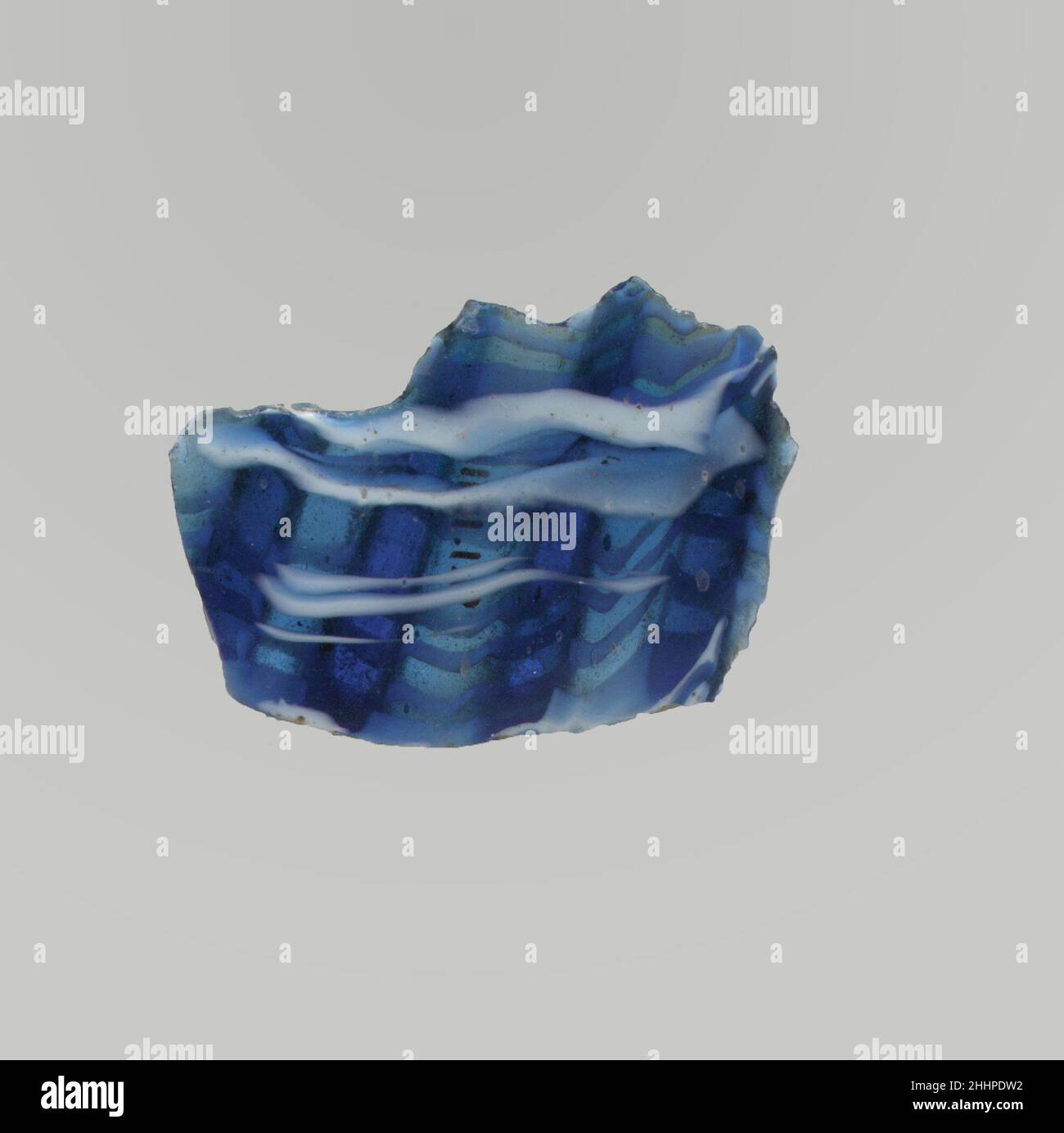 Glasmosaik geripptes Schalenfragment Ende des 1st. Jahrhunderts v. Chr.–Anfang des 1st. Jahrhunderts n. Chr. römisches Körperfragment.Durchscheinendes Kobaltblau und opakes Weiß.leicht gewölbte, nach unten gewölbte Seite.Mosaikmuster aus Bändern, die aus Längen eines einzelnen Rohres in einem blauen Boden mit welligen, horizontalen Bändern aus weißen Fäden in parallelen Linien gebildet werden; Außen, Teil von vier vertikalen Rippen mit abgerundeten Außenflächen, nach unten verjüngend.Poliertes Innenleben; Lochfraß von Oberflächenblasen im Inneren; stumpfes, Lochfraß und schwache Witterung an der Außenseite und gezackte Kanten. Glasmosaik gerippte Schale Fragment. Römisch. Ende 1st Jahrhundert B. Stockfoto