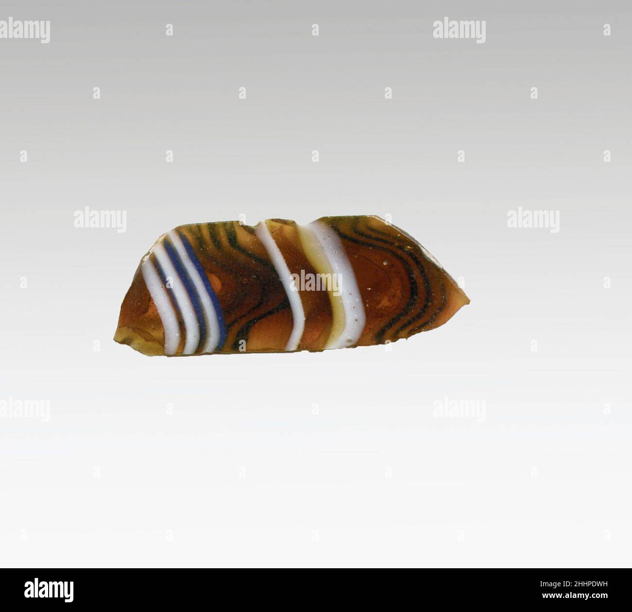 Mosaikglas geripptes Schalenfragment Ende des 1st. Jahrhunderts v. Chr.–Anfang des 1st. Jahrhunderts n. Chr. römisches Körperfragment.Durchscheinendes honigbraun, kobaltblau und opakes Weiß.konvex geschwungene Seite, die sich nach unten verjüngt.Mosaikmuster aus Abschnitten eines einzelnen Rohres in braunem Boden mit unregelmäßigen welligen weißen und blauen Fäden in parallelen Linien gebildet; Außen, einzelne lange vertikale Rippe mit abgeflachte Oberseite und abgerundeter Außenkante. Innen poliert; innen mit Lochfraß von Oberflächenblasen; außen stumpfend und cremig schillernd; außen etwas verwittert und zerklüftete Kanten abgesplittert. Mosaikglas gerippte Schale fragm Stockfoto