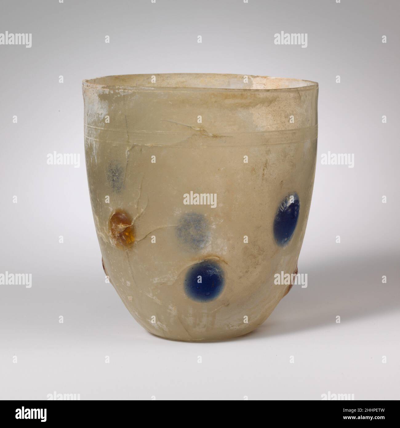 Glasbecher 4th. Jahrhundert n. Chr. römisch, rheinisch farblos; transluzente kobaltblaue und honiggelbe Flecken.unebener vertikaler Rand, abgerissen und geschliffen, leicht ausgespielt; hohe, konvexe Seite zum Körper, nach unten verjüngend; konkaver Boden.unmittelbar unter dem Rand eine einzelne, mit Rädern geschnittene horizontale Linie; Unten ein Band, bestehend aus zwei radgeschnittenen horizontalen Linien mit Rad-abraspelten Linien zwischen ihnen; um den mittleren und unteren Teil der Seite ein Band von aufgebrachten Blobs, bestehend aus zwei unregelmäßigen horizontalen Linien von vier abwechselnd Sätze von zwei Blobs der gleichen color.Complete, aber viele interne Risse; ein paar Nadelstich und Stockfoto