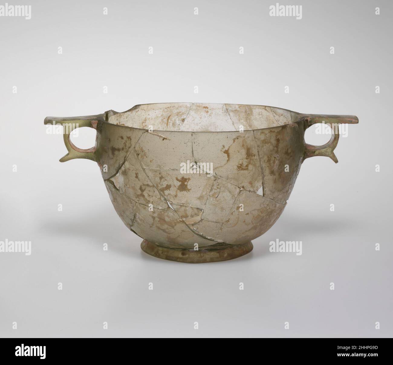 Glas-Skyphos (Trinkbecher) Ende 3rd–Anfang 2nd Jahrhundert v. Chr. griechisch farblos mit blassgrünlichem Farbton. Leicht umgekehrter Rand, mit oberer Kante flach geschliffen; konvex geschwungene Seite nach unten verjüngend; ausgespriebener Basisring mit abgerundeter unterer Kante; leicht wellig, flacher Boden; An den Seiten des Körpers wurden zwei Ringgriffe angebracht, die aus Rohlingen herausgeschnitten wurden, die von erhöhten quadratischen Stellen umgeben sind, mit flachen Daumenstützen über Ringen und hervorstehenden Flügeln über und unter.Körper gebrochen und repariert, mit einem großen Span im Rand und mehreren Löchern im Körper, aber beide Griffe intakt; Nadelstich und größere Blasen; Dulling, pitting, pa Stockfoto