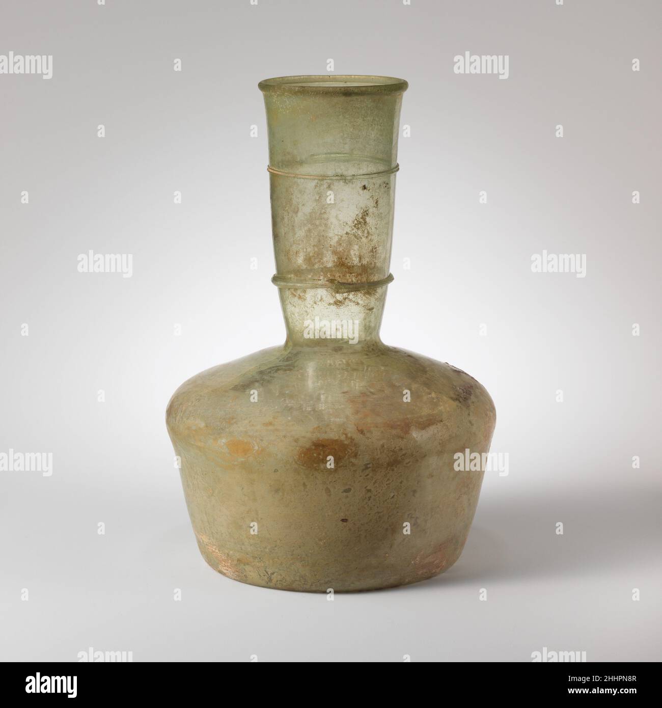 Glasflasche 5th. Jahrhundert n. Chr. oder später römisch Durchscheinend hellblaugrün; Spuren in gleicher Farbe. Umgeknickter Rand, über- und eingefaltet; zylindrischer Hals, nach unten zugespitzt mit horizontaler Einrückung an der Basis; breite, schräge Schulter mit abgerundeter, leicht auskragender Außenkante; zylindrischer Körper mit nach unten zulaufenden Seiten; Tief eingeschobene Unterseite.am Hals eine feine Spur, als längliche Unterlage aufgetragen und zweimal um zwei parallele Linien gewickelt; eine zweite Spur, weiter unten am Hals, umwickelt als eine dickere überlappende Linie.intakt; einige Blasen und schwarze Verunreinigungen, vor allem in Hals und Rand, und zahlreiche Brandung Stockfoto