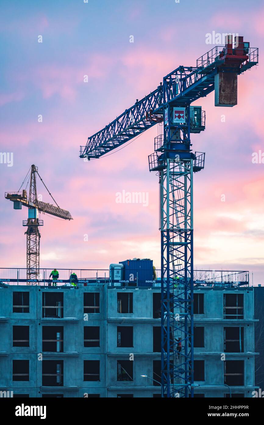 Baustelle mit Kranen, Neubauten oder im Bau befindlichen Häusern mit Arbeitern mit Helm bei der Arbeit und buntem Himmel im Hintergrund, vertikal Stockfoto