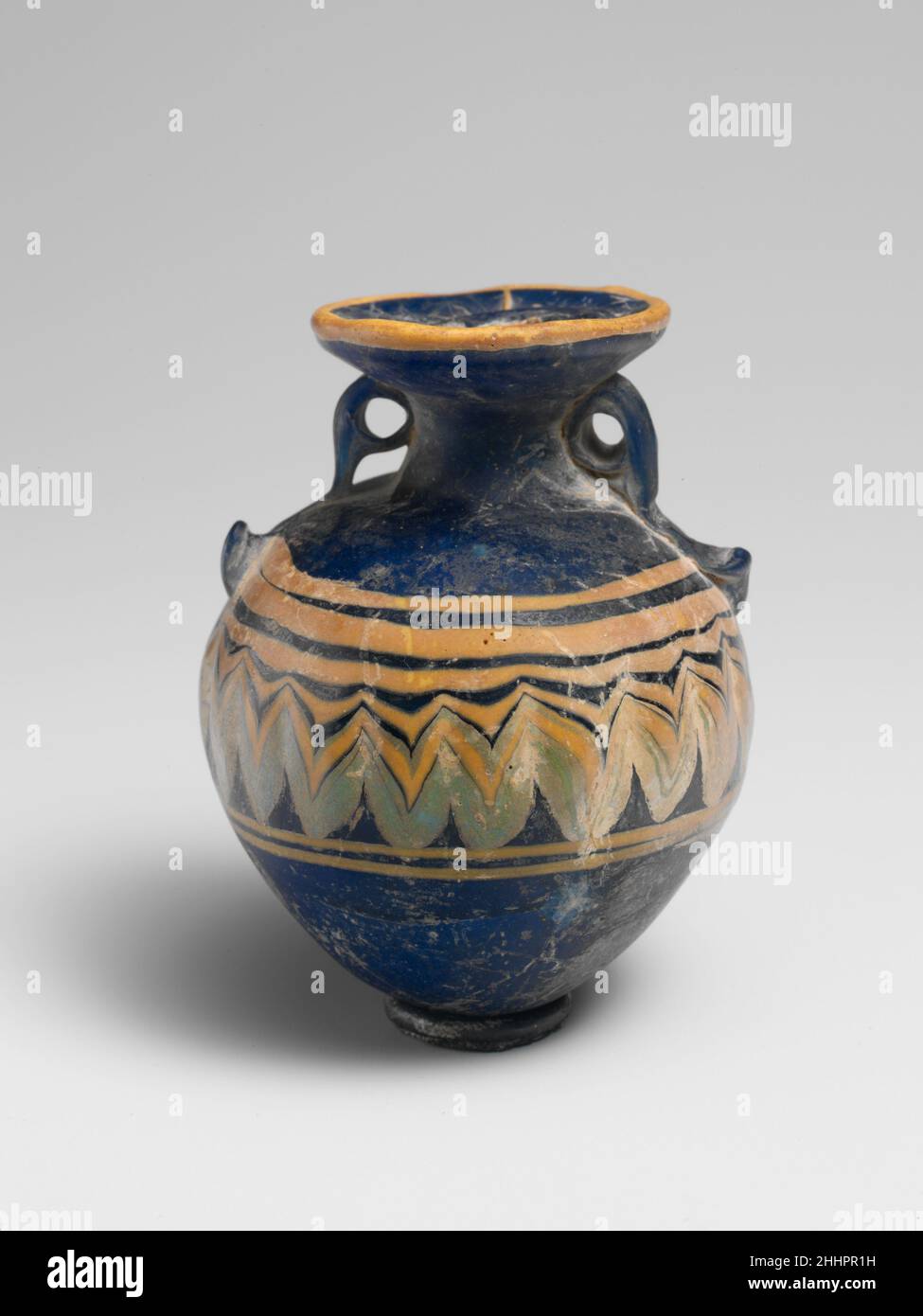 Glas Aryballos (Parfümflasche) Ende des 6th.–5th. Jahrhunderts v. Chr. Griechisch, östliches Mittelmeer Durchscheinendes Kobaltblau, mit Griffen gleicher Farbe; Trails in opak-gelb und opak-türkisblau. Breite nach innen geneigte Randscheibe mit unebener Oberfläche; kurzer konkaver Hals; unebene, eckige Schulter; fast kugelförmiger Körper; konvexer, spitz zulaufender Boden; Zwei große Ringgriffe mit geklopften Schwänzen, die über der Traildekoration angebracht sind, erstrecken sich von Schulter zu Hals und Unterseite der Randscheibe.Eine gelbe Spur wird am äußeren Rand der Randscheibe angebracht; eine weitere gelbe Spur wird am äußeren Rand der Schulter angebracht und spiralförmig nach unten gewickelt, bei Stockfoto