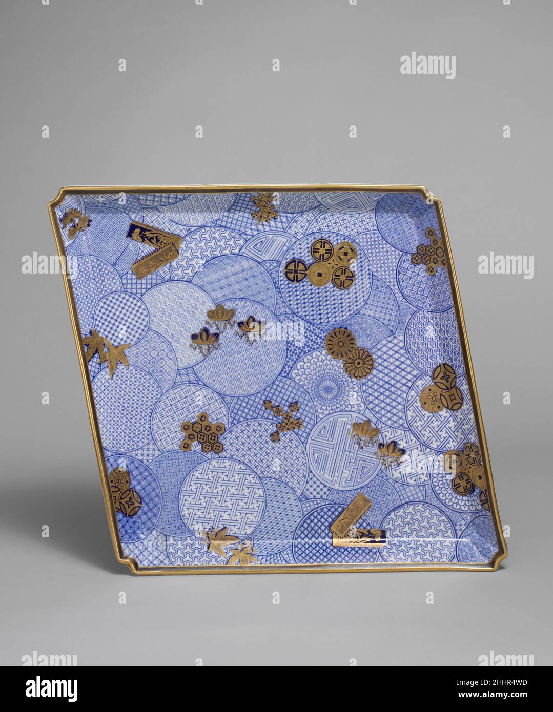 Tablett 1884 Worcester-Fabrik das Ranzenförmige Tablett ist in Unterglasur-Blau mit einem komplexen Design aus Kreisen bedruckt, die mit unterschiedlichen Bodenmustern gefüllt sind, als wären zahlreiche verzierte Scheiben über das Tablett verteilt worden, überlappen und überlappen sich von ihren Nachbarn. Über allen sind Klumpen von Schablonenmotiven in Gold, bestehend aus verzierten Kreisen, Quadraten, Sechsecken, Pflanzen, Blumen, Stiftboxen und Blätter. Die Marke S.Y. in Gold auf der Unterseite des Tabletts ist die eines anonymen Vergolders. Andere Marken sind eine gedruckte Royal Worcester Company Marke, ein V für das Jahr I884 und die des Unternehmens Stockfoto