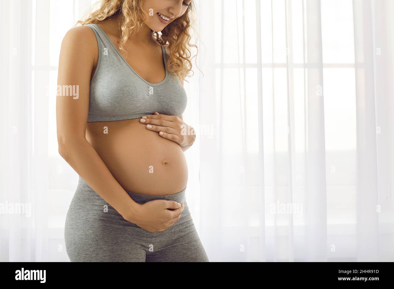 Glückliche Frau, die ein Baby erwartet, das zu Hause am Fenster steht und ihren Bauch berührt Stockfoto