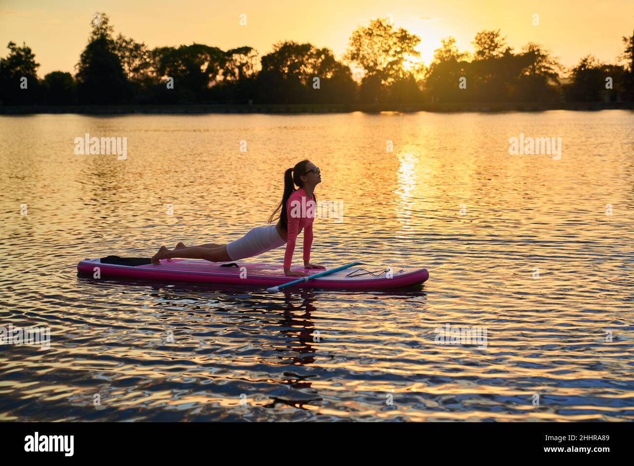 Junge athletische Frau in Sportkleidung, die während des Sonnenuntergangs über dem See sup Yoga praktiziert. Kaukasische Brünette, die Freizeit für Outdoor-Training verbringt. Lieblingshobby-Konzept. Stockfoto