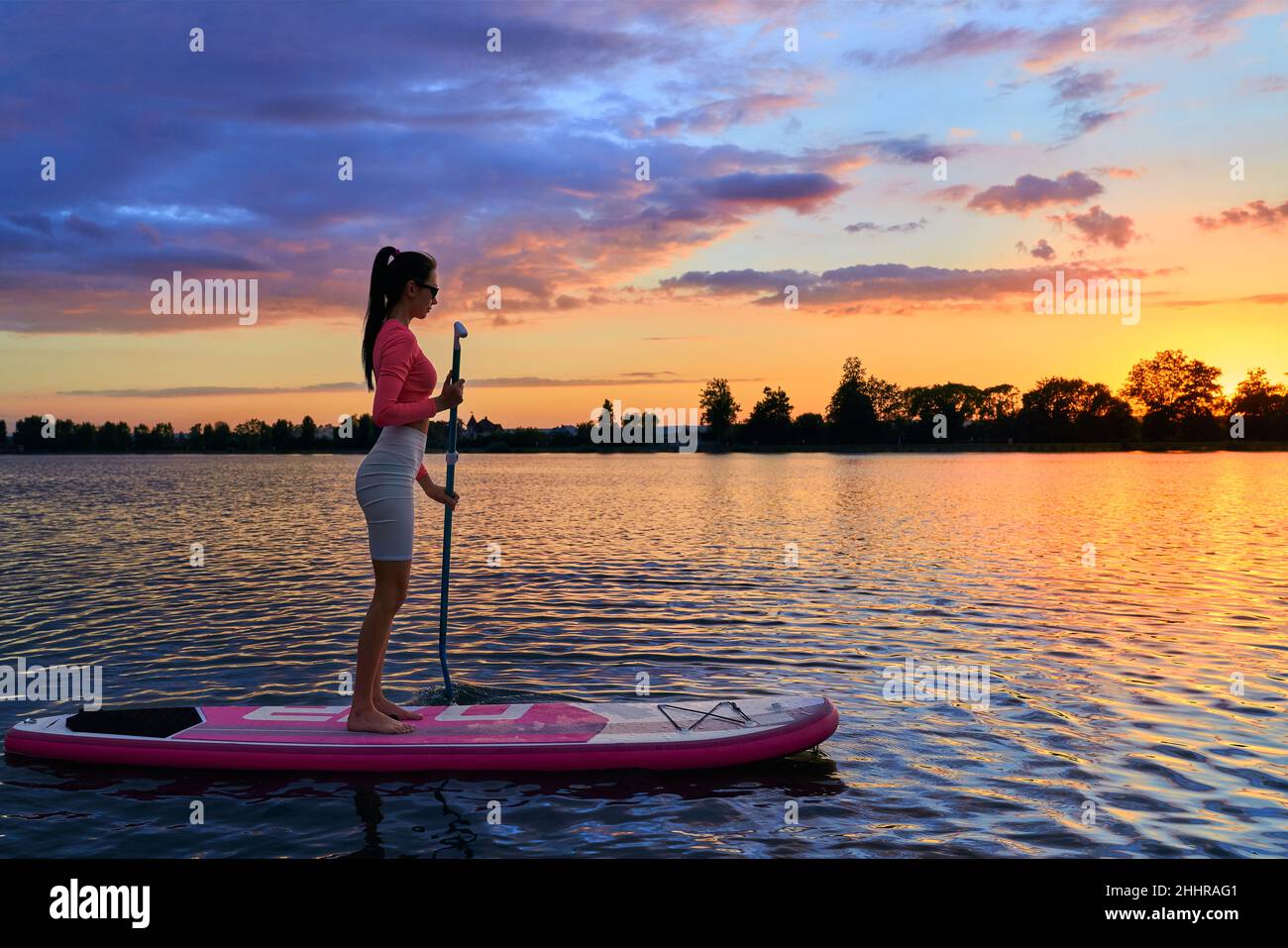 Kaukasische athletische Frau schwimmt auf dem Brett mit langem Paddel in den Händen. Junge kaukasische Brünette Workout auf Natur während schönen Sonnenuntergang. Konzept für einen gesunden Lebensstil. Stockfoto