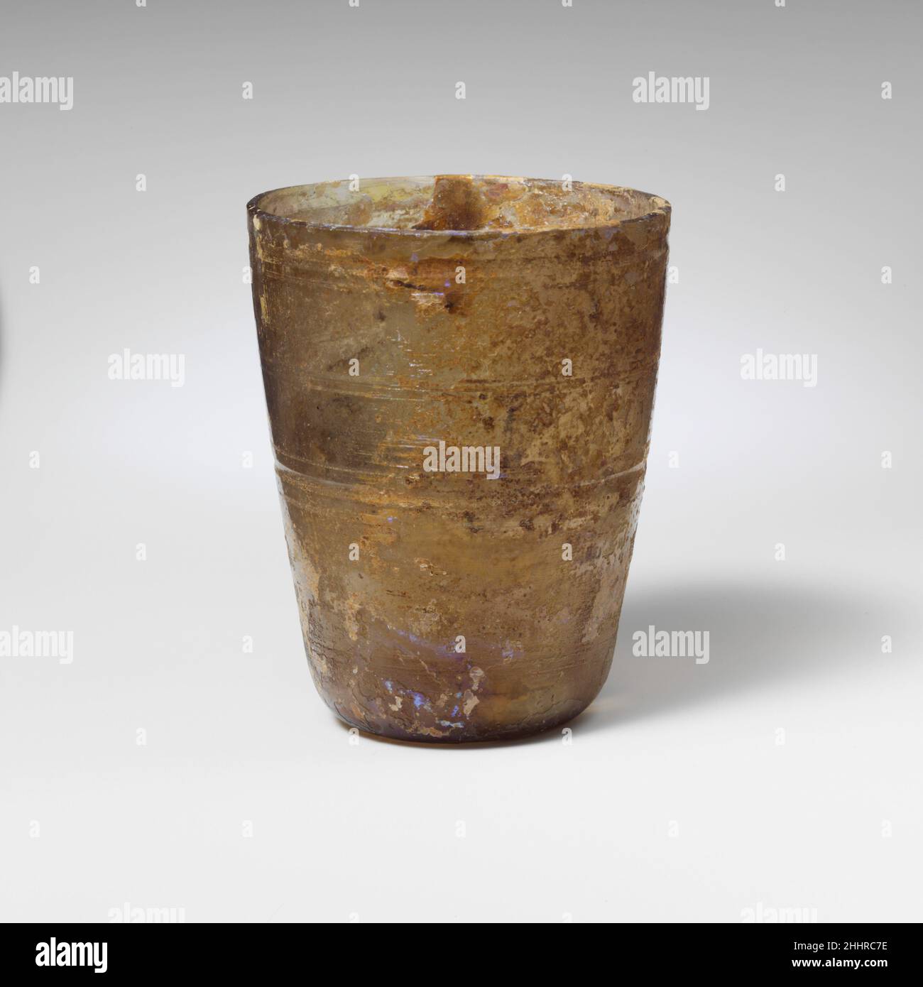 Glasbecher 1st. Jahrhundert n. Chr. römisch farblos mit violetten Streifen unter dem Rand.abgeklopfter Rand; fast gerade Seiten zum Körper, nach unten verjüngend; konkaver Boden. Dickwandig.Radschnitt-Dekoration auf dem Körper in vier Bändern, bestehend aus einer tiefen Nut und schwachen Linien unter dem Rand; zwei parallele Linien; eine tiefe Nut, die über und unter einer einzigen Linie faltet; und zwei weitere parallele Linien nahe unten.intakt; Nadelstich-Blasen; Flecken von Abstumpfen, Lochfraß und irisierender Verwitterung. Glasbecher 239838 Stockfoto