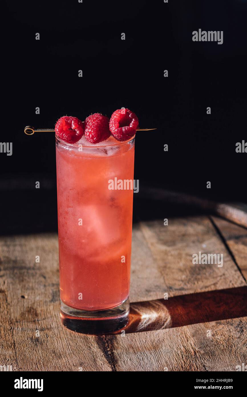 Hohes rosafarbenes Cocktailgetränk mit drei Himbeeren auf Cocktailpickel, auf Holzfass Stockfoto