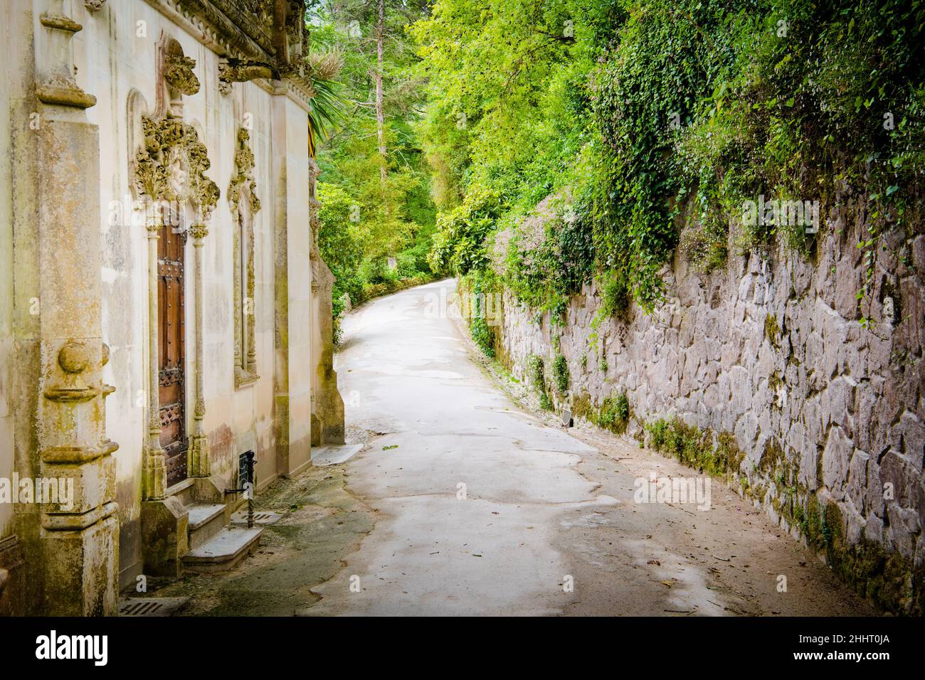 Ein Pfad schlängelt sich durch einen üppigen grünen Garten, vorbei an einem wunderschönen weißen Gebäude und einer mit Efeu bedeckten Steinmauer in Sintra, Portugal Stockfoto
