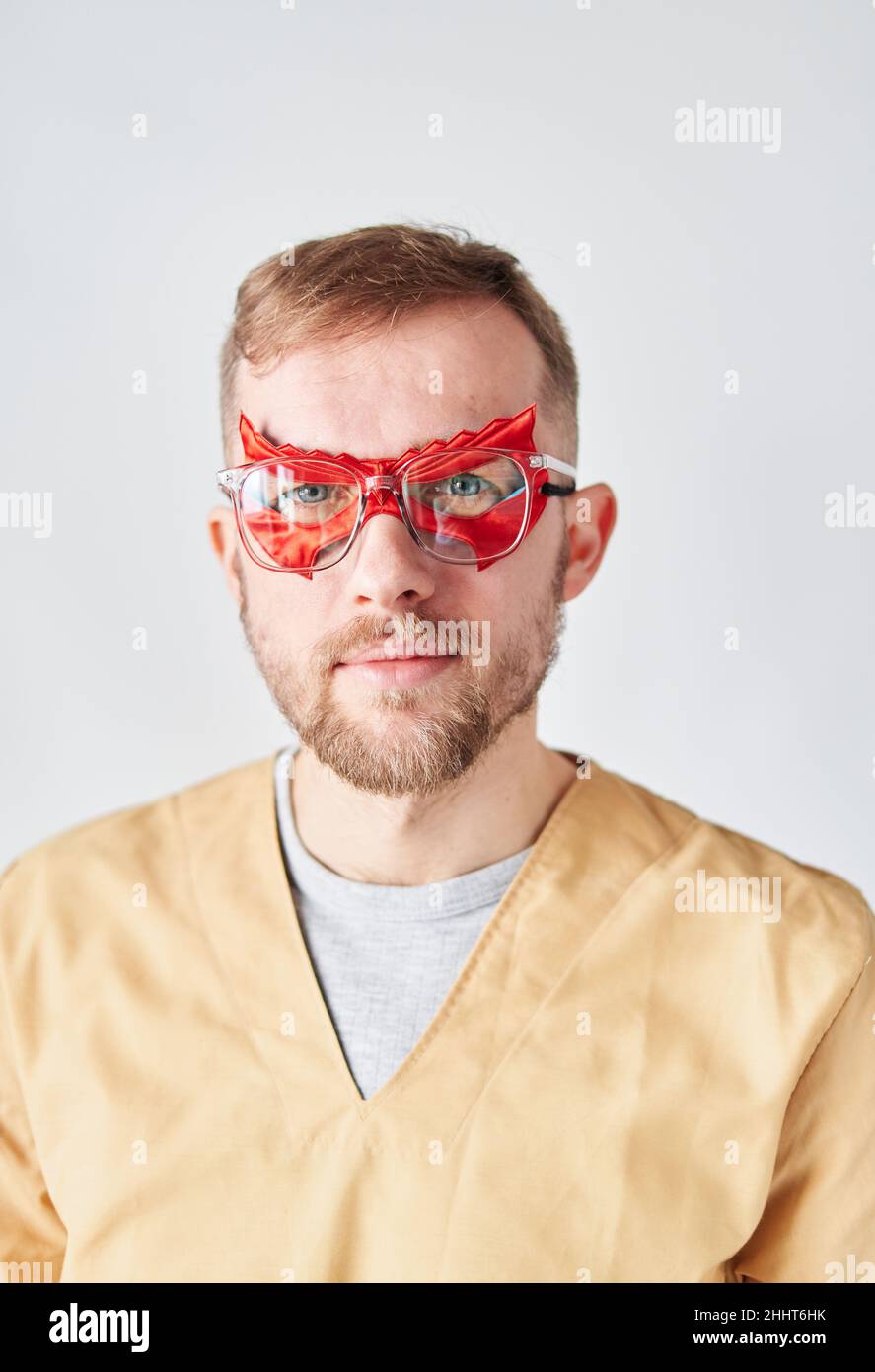 Nahaufnahme des Porträts eines männlichen Supermannes, eines Arztes oder einer Krankenschwester. Porträt eines hübschen kaukasischen männlichen Arztes, der eine Superheldenmaske mit rotem Gesicht, eine Brille und eine beigefarbene medizinische Uniform trägt. Hochwertige vertikale Fotos Stockfoto