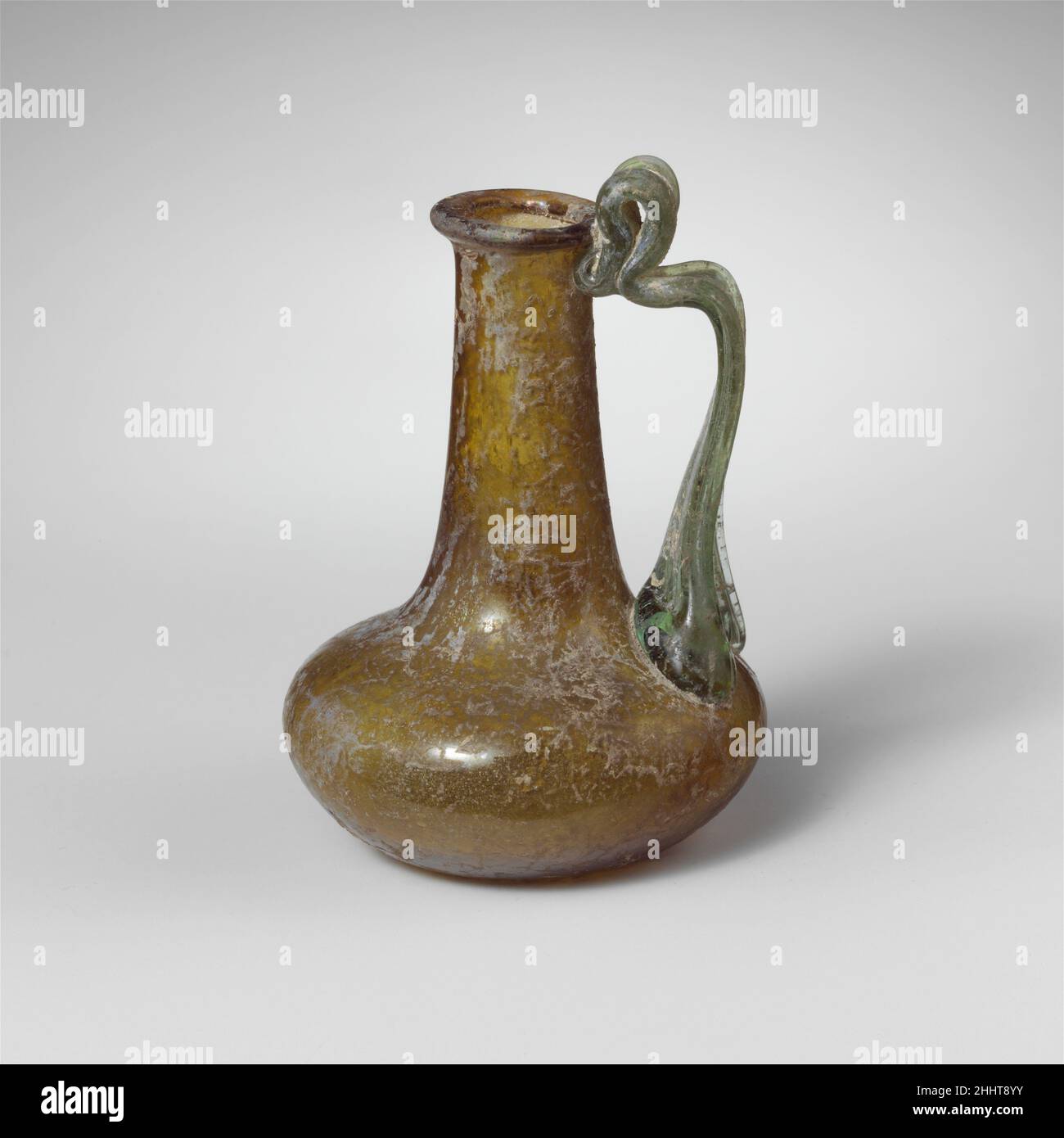 Glaskanne 1st–2nd century A.D. römisch Durchscheinend Gelbbraun, mit lichtdurchlässigem hellgrünem Griff.Rand ausgeklappt, über und in, mit abgeflachte obere Oberfläche; zylindrischer Hals, sich nach unten ausdehnend; hockiger, bulböser Körper; Konkaver Boden; breiter Griff mit Riemen, der in einem großen Pad mit zwei äußeren Krallen am oberen Teil des Körpers angebracht wird, gezogen, dann vertikal gedreht und am Hals unter dem Rand mit vertikaler Schlaufe als Daumenstütze oben befestigt. Intakt; einige Nadelstich-Blasen; Dullende und schwach kalkige Verwitterung mit etwas Schillern, Bodenverkrustungen und weißlich schillernder Verwitterung im Innenraum. Glaskanne 244661 Stockfoto