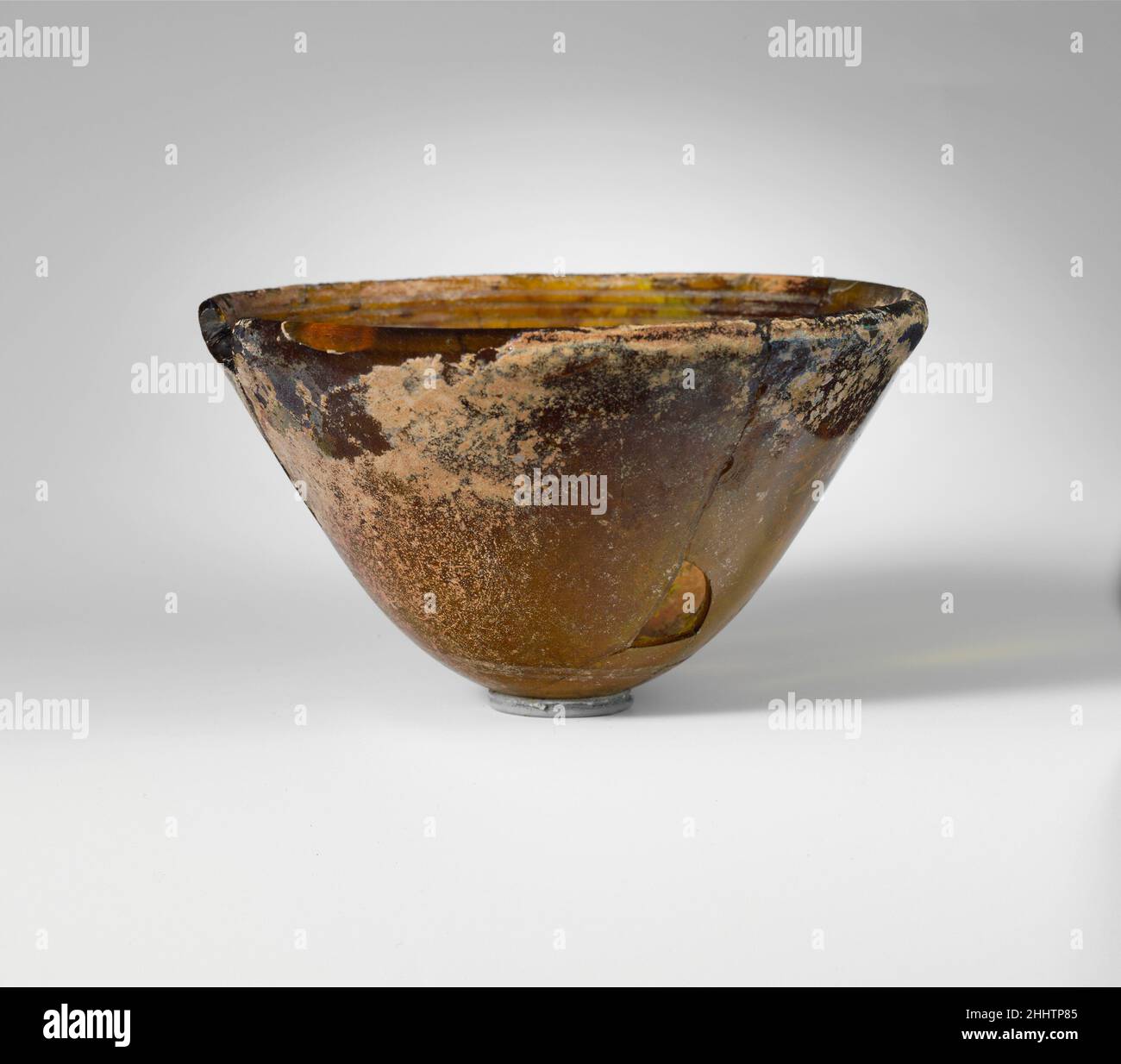 Konische Glasschale Mitte–2nd bis Anfang 1st Jahrhundert v. Chr. griechisch Transluzent honigbraun.aufrecht abgerundeter Rand mit fast spitz zulaufender Oberkante; gerade Seite, schräg nach unten verjüngend; konvexer Boden.im Inneren drei horizontale Rillen in einem Band unterhalb des Randes geschnitten; Außen zwei leicht abrassierte Rillen in einem Band um den Unterkörper.gebrochen und repariert, mit zwei Löchern in der Seite und großem Span im Rand; wenige Blasen; Flecken von tiefem Lochfraß und Verwitterung mit brillantem Schillern auf der Außenseite, und dicke cremige Verwitterung auf der Innenseite.. Konische Glasschale 245789 Stockfoto