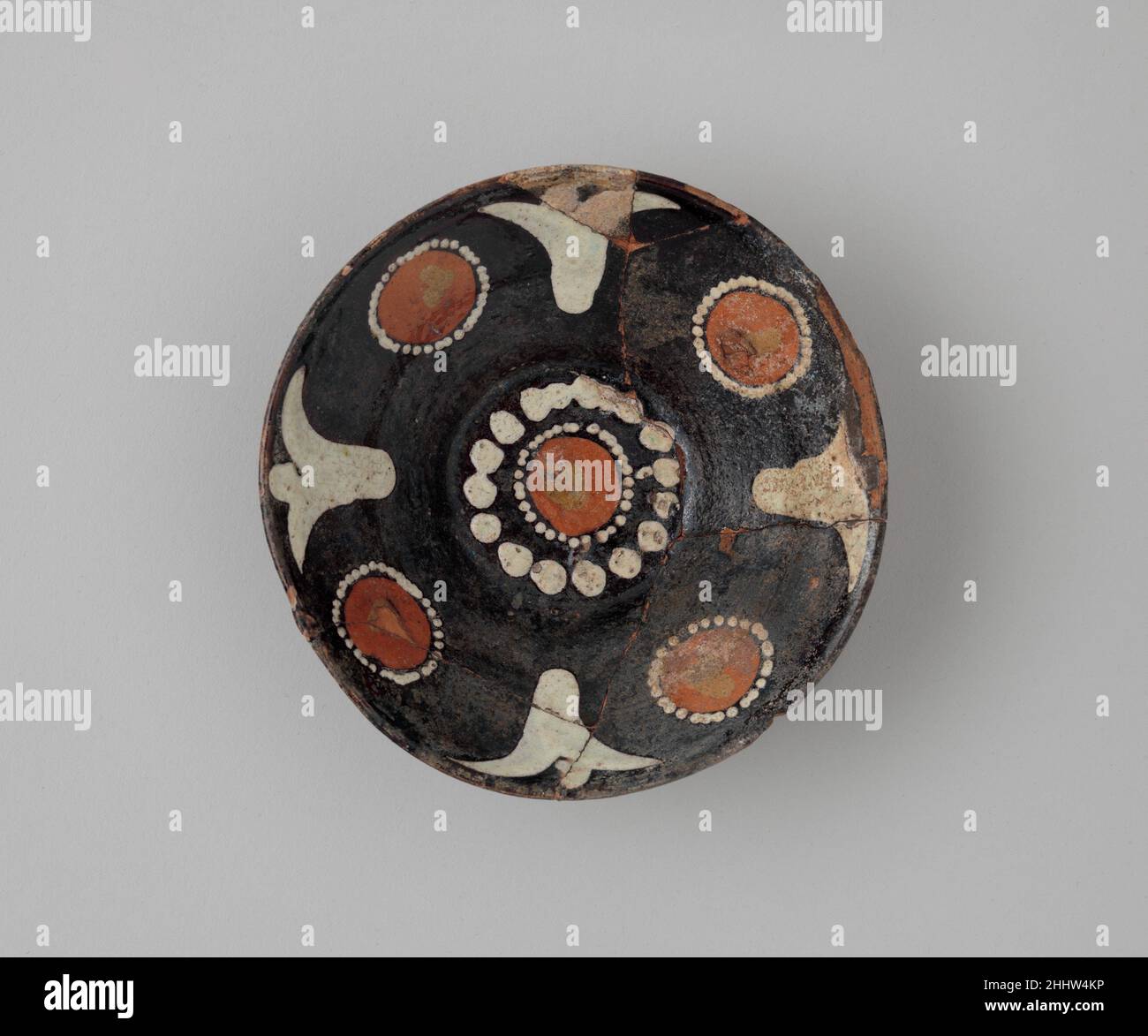 Schale mit Polychromdeko auf schwarzem Rutschboden 10th Jahrhundert Schalen aus kühn gefärbten Slips (verdünnter Ton) wurden in der gesamten Umgebung von Nishapur hergestellt. Diese Slip-Ware unterscheidet sich leicht von der Keramik, die im Rest des abbasidischen Reiches hergestellt wurde, und steht in keinem Zusammenhang mit der populären chinesischen Keramik, die auch in Nishapur hergestellt wird. Ähnliche Rosetten schmücken Schalen von anderen nahe gelegenen Orten, einschließlich Samarqand, aber die eigentümliche Blattgestaltung ist häufiger bei den Funden aus Nishapur. Diese kleine Schüssel wurde wahrscheinlich für den täglichen Gebrauch im Haushalt gemacht. Schale mit Polychrom Dekorieren Stockfoto